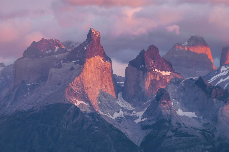 Pink and purple sunlight is cast over the spiky mountain peaks of Cuernos del Paine at Torres del Paine National Park, Chile.